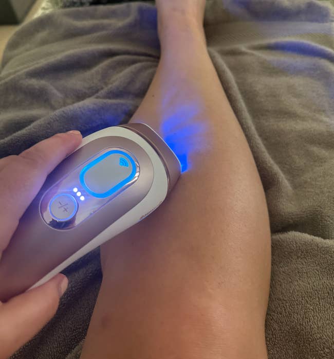 Close-up of a handheld device being used on a person's leg, possibly for hair removal or skin treatment. The device emits a blue light