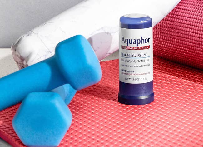 Aquaphor healing balm stick on a red yoga mat with blue dumbbells and a water bottle nearby. Perfect for post-workout skin care