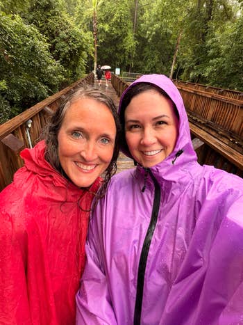 Two people smiling in rain gear on a wooded path, enjoying a rainy day
