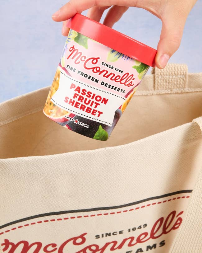 A hand places a container of McConnell's Passion Fruit Sherbet into a branded tote bag