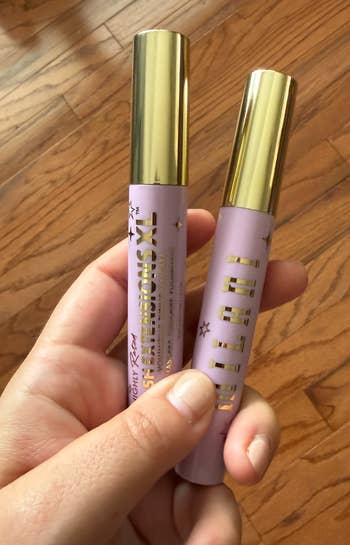 A hand holding two Milani makeup products with golden caps, likely mascaras, against a wooden floor background