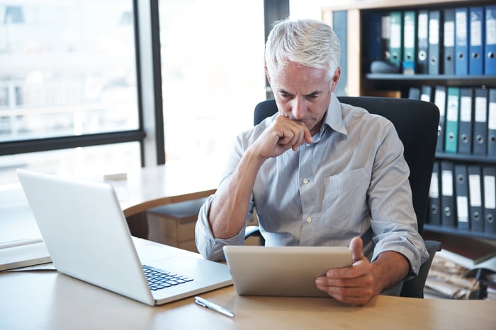 Person sitting at a desk with a laptop open, looking at a tablet.