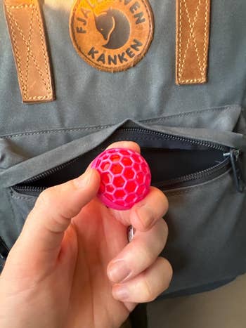 Hand holding a small, textured pink ball in front of an open backpack pocket