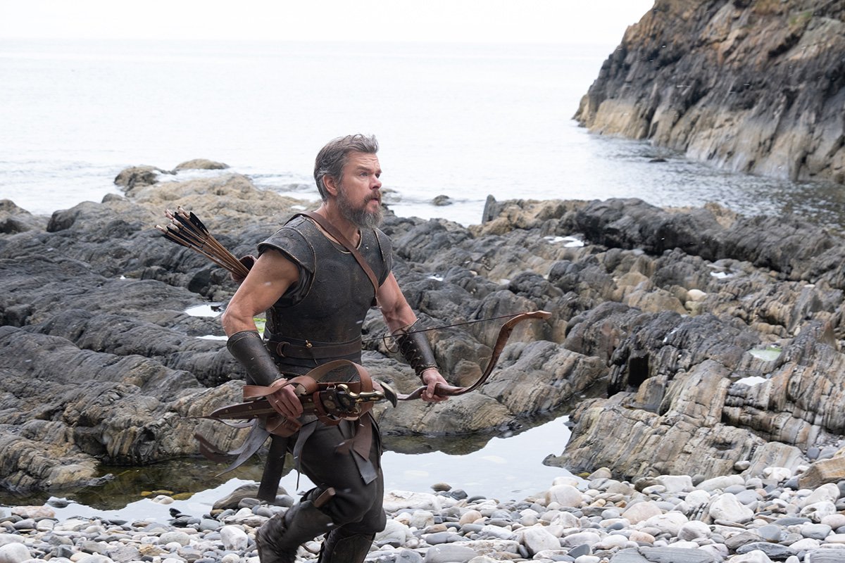 Matt Damon as Odysseus holding a bow and arrow and running up a rocky beach in The Odyssey