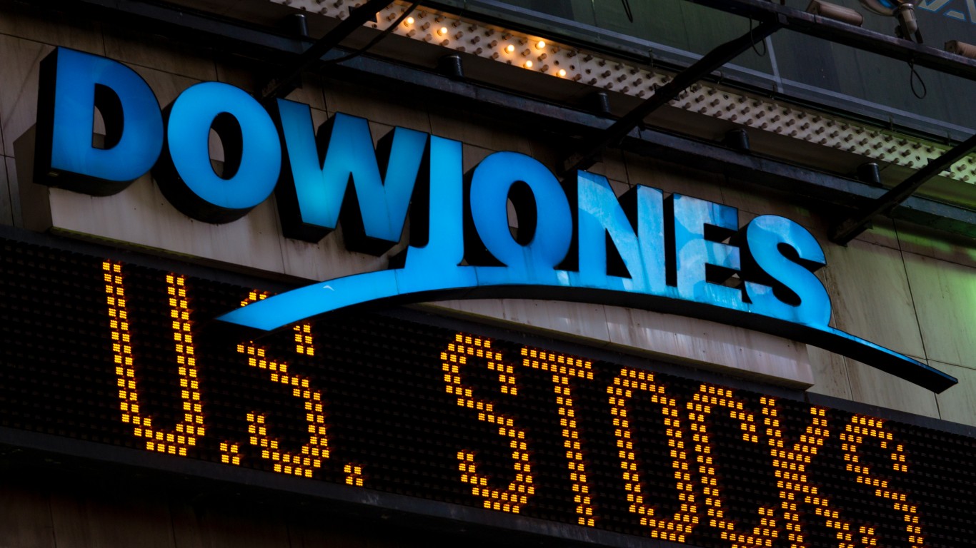 A nighttime close-up shot of the Dow Jones sign with bright blue block letters 'DOWJONES' standing out against a dark building facade. Below the sign, an LED ticker displays the words 'US STOCKS' in glowing orange dot matrix letters, partially obscured on the right by a blue swoosh design extending from the main logo. The image is taken slightly from below, looking up at the illuminated signs.