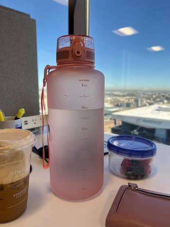 Large frosted water bottle with measurement markings on an office desk, alongside a coffee cup, food container, and wallet, against a city view