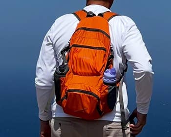 Person overlooking the ocean, wearing a white cap, long-sleeve shirt, shorts, and an orange backpack