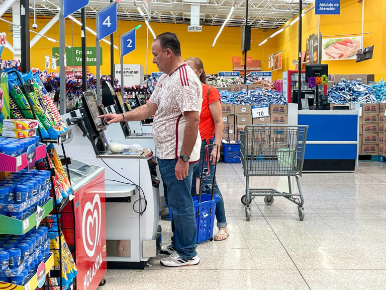 Customer using a self-checkout kiosk at a Walmart store, with shopping carts and checkout lanes in the background.