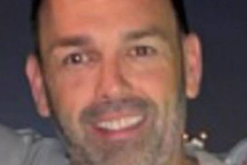 A close-up portrait of an individual with a clean-shaven face, sporting a warm, broad smile and exuding an air of contentment. The person is dressed in a gray shirt, and their expression is framed by short, neatly trimmed hair.