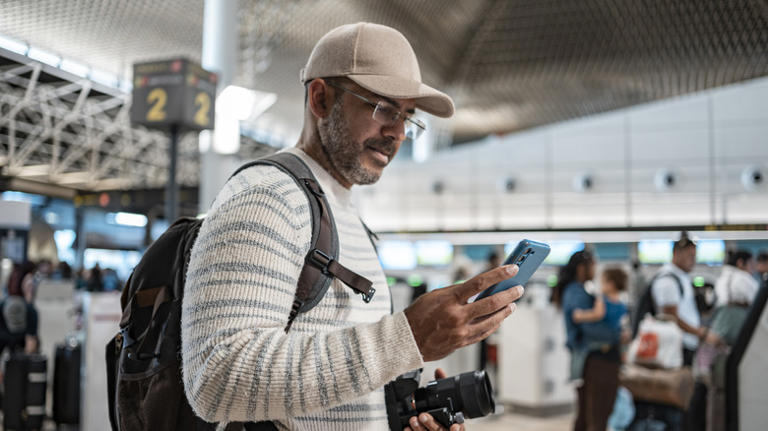 Middle aged man wearing a ball cap using his cell phone at the airport getting ready for a trip