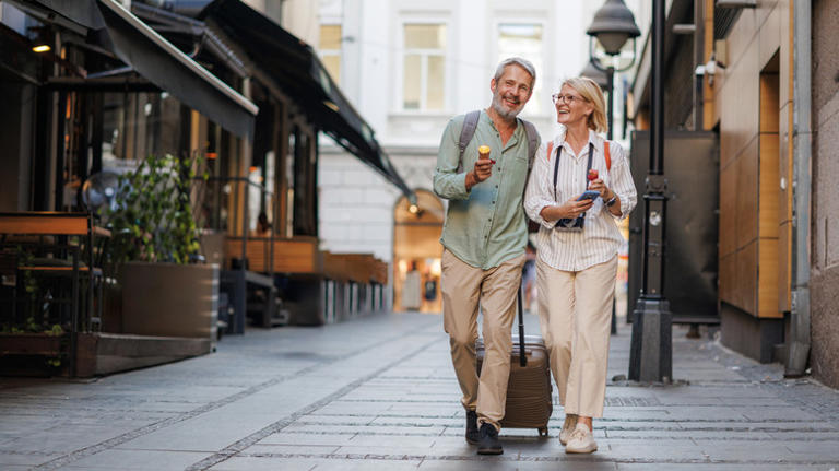 Senior couple with suitcase and backpacks strolling a European city street, sharing an ice cream cone and laughing