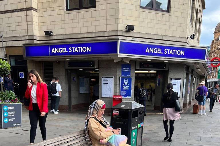 A woman wearing a veil and a long overall sits on a wooden bench busy with her mobile phone outside Angel tube station