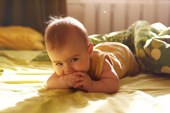 A baby with light brown hair lies on a bed, smiling and chewing on a hand. The room is softly lit with yellow and green bedding.
