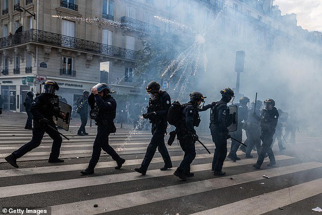 Police officers carrying batons and shields are seen rushing along a Parisian street as a firework explodes above them