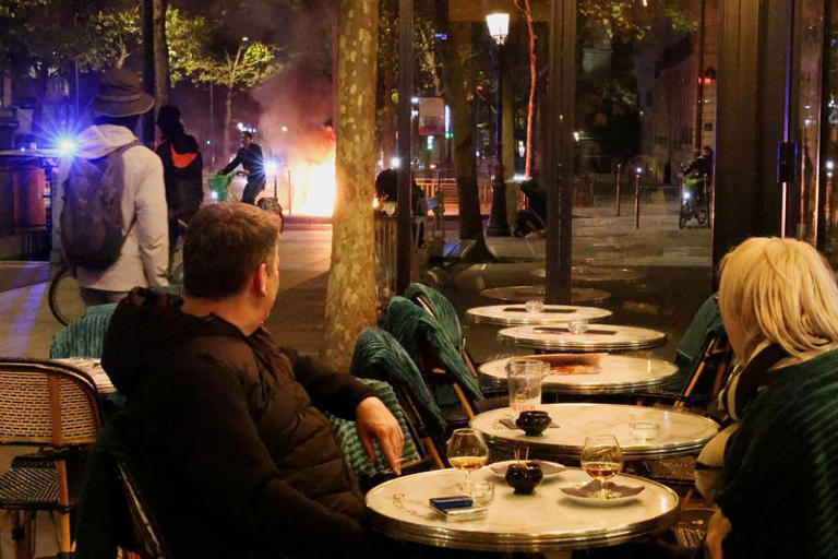 Customers at a cafe watch a fire set by rioters at the end of a day of nationwide cost-of-living protests in Paris, France, October 17, 2026. REUTERS/Yann Tessier.
