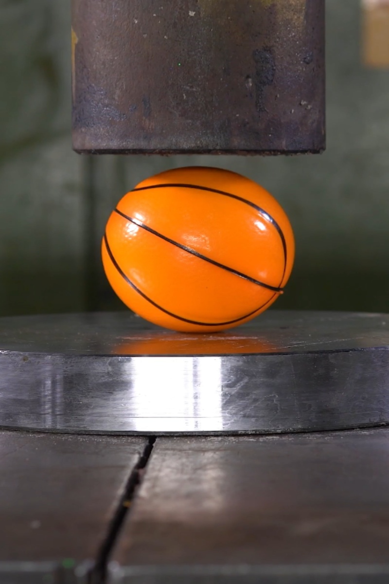 Prensa hidráulica aplastando un balón de baloncesto de color naranja