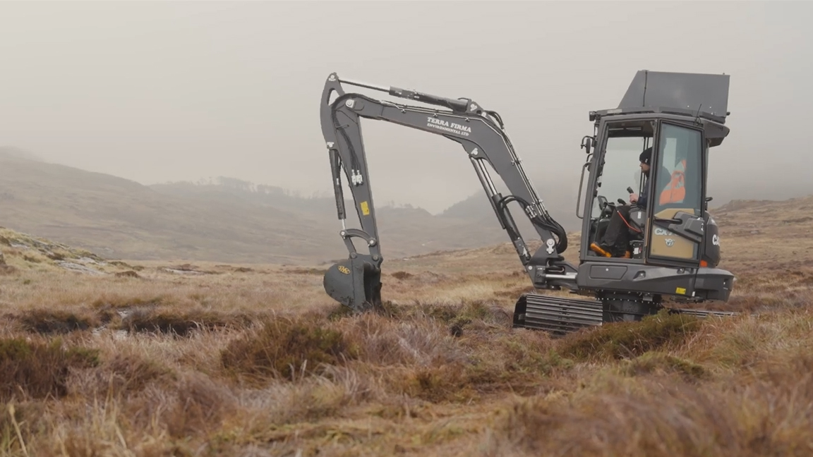 Mini excavator digging through thick grass on open moorland