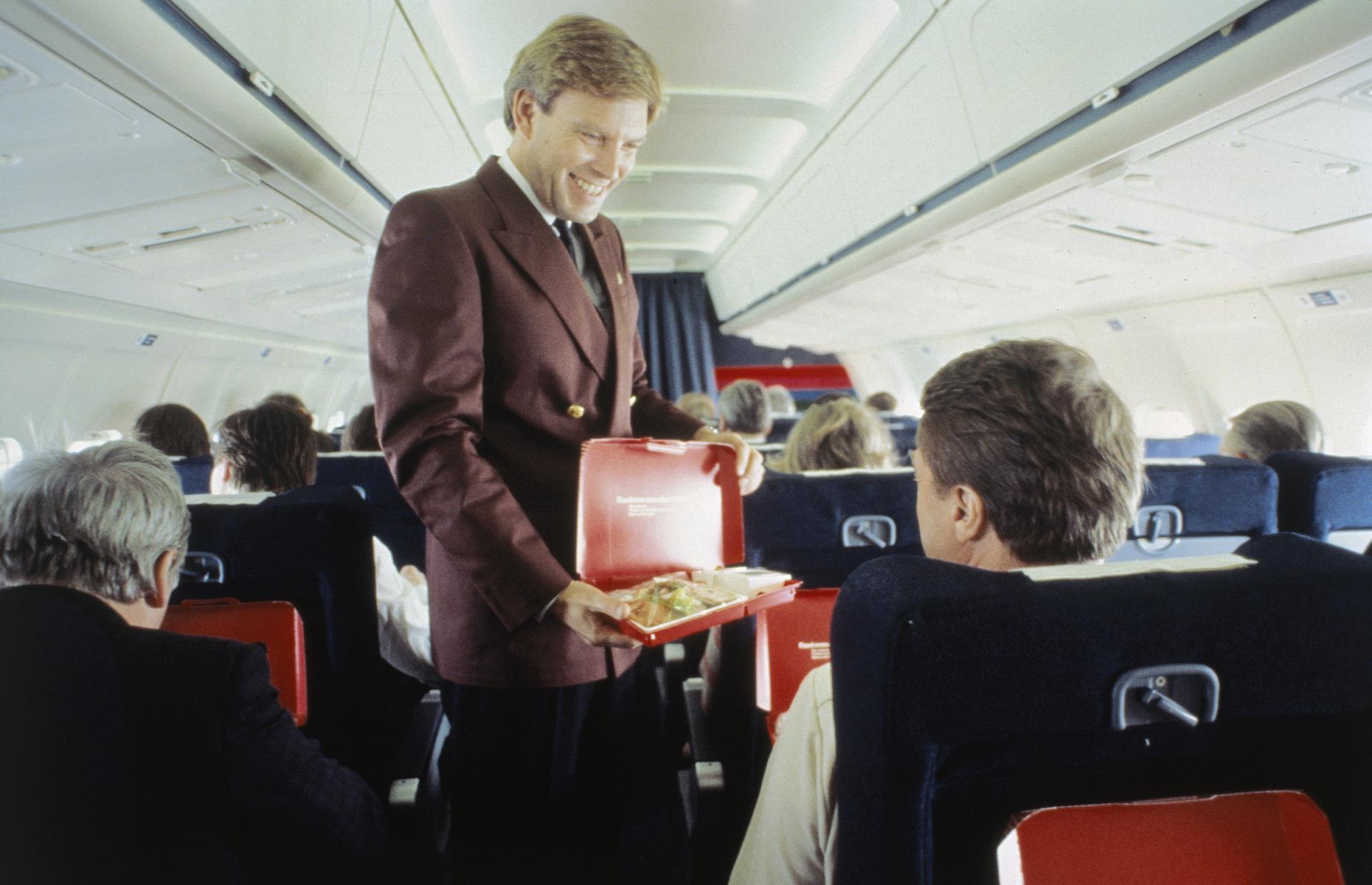 Slide 40 of 51: Through this decade, as flying became more and more commonplace, the economy class cabin looked much as it does today. Lavish, multi-course meals had been mostly replaced with more humble dinners served from boxes or trays. This photo shows an SAS flight attendant serving boxed meals to passengers.