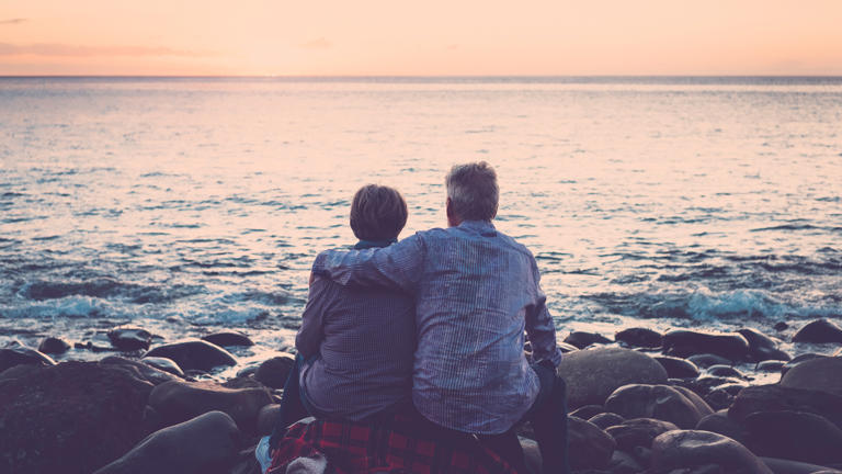 love and romance with adult matures – elderly couple sitting and hugging each other looking at the sea at sunset relaxing. Concept of vacation, leisure time, relaxation –