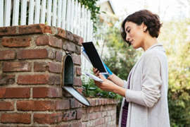 a person standing in front of a brick building: Woman sorting through mail at her mailbox.