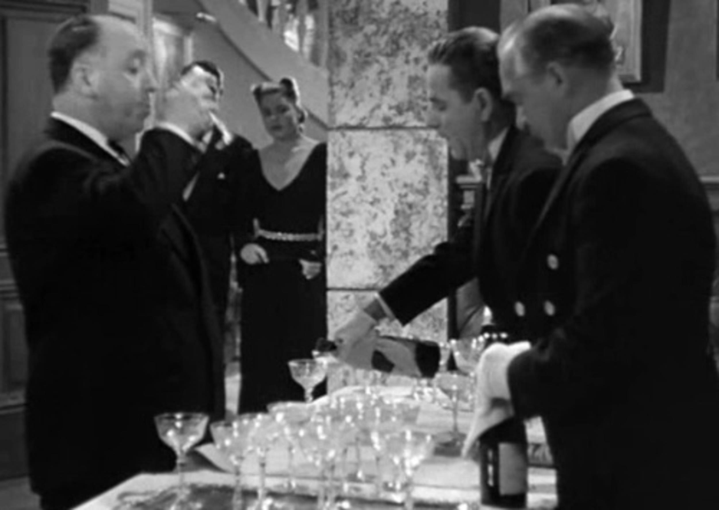 <p>A crowded elevator is an easy place for a cameo. So is a lavish party. You can see Hitchcock front-and-center at Claude Rains’ extravagant get-together having a glass of champagne filled. Seems like he’s having a nice time.</p>