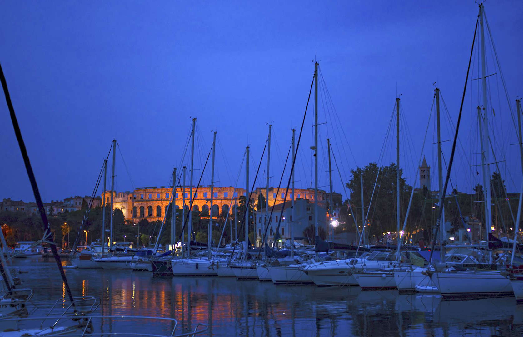 Slide 19 of 41: In this present-day night shot, Pula Arena is juxtaposed with its more modern surroundings. The harbour is crammed with sleek sail boats and lights glitter on the water at dusk. Today history buffs can explore the amphitheatre and it also typically hosts concerts, dance shows and festivals in place of bloody gladiatorial battles.