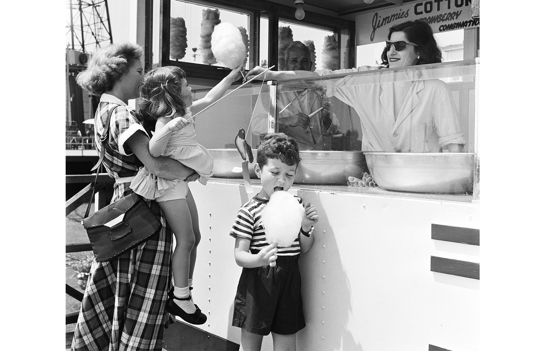 Coney Island attracted holidaymakers with its bounty of sweet treats too. In this gloriously nostalgic snap, a mother lifts up an eager child, so she can accept a sugary cloud from Jimmies cotton candy shack.