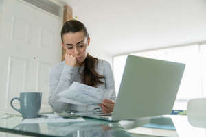 a woman sitting at a table using a laptop computer: Woman paying her utility bills online and looking worried while staying at home during the pandemic - home finances concepts.