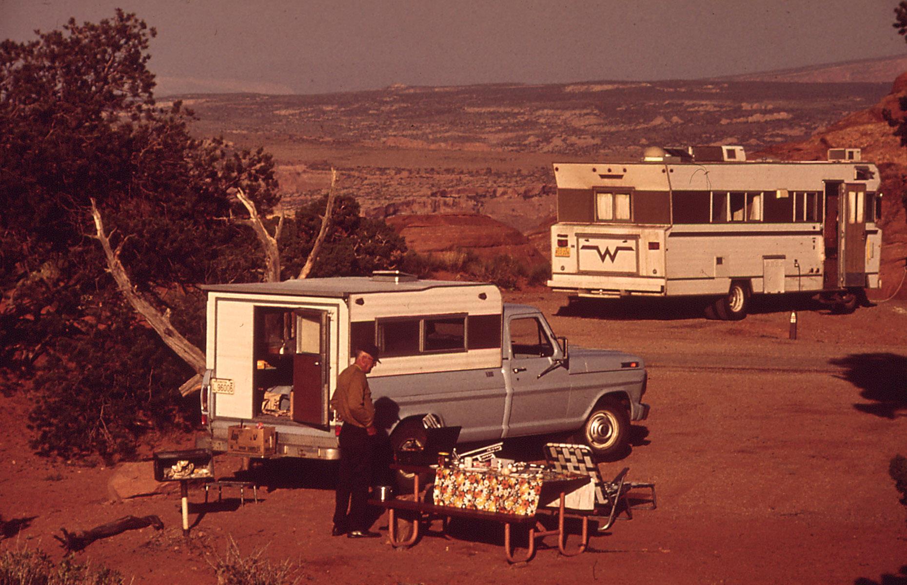 This snap shows a Winnebago motorhome in its natural habitat: in America's beautiful backyard with far-reaching views out across the countryside. The red-tinged photo captures Arches National Park, Utah in the Seventies, with the folks in the foreground setting up for an alfresco dinner.