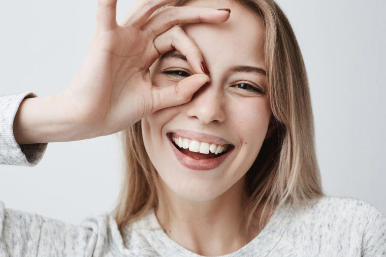 Close up portrait of beautiful joyful blonde Caucasian female smiling, demonstrating white teeth, looking at the camera through fingers in okay gesture. Face expressions, emotions, and body language