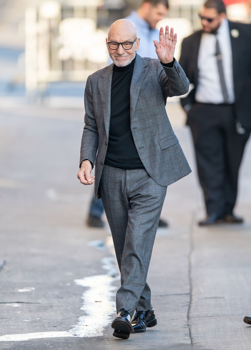 <p>Sir Patrick Stewart waves while arriving for a Nov. 12, 2019 appearance on ‘Jimmy Kimmel Live!’ He wore a grey suit with a subtle brown grid on it.</p>