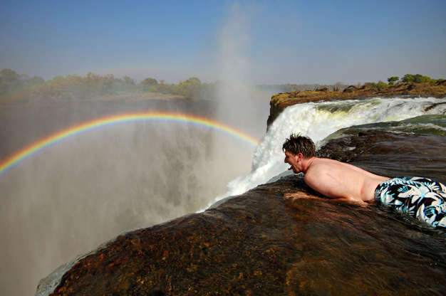 ÎÎ¹Î±ÏÎ¬Î½ÎµÎ¹Î± 20 Î±ÏÏ 24: Man lying on rock with rainbow and waterfall, Devil's pool, Victoria Falls, Zambia.