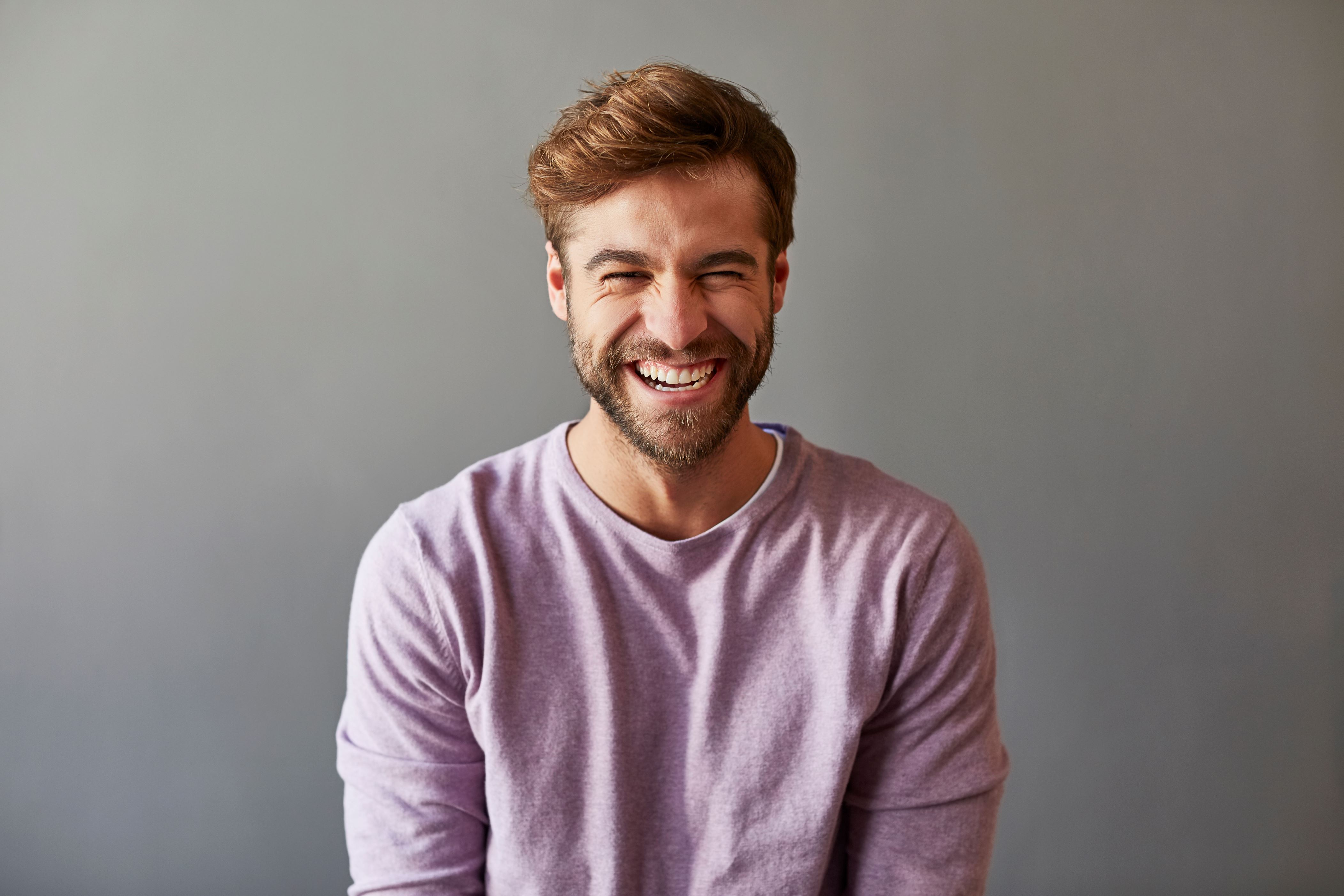 Slide 18 de 21: Male professional laughing over gray background. Cheerful businessman is wearing purple t-shirt.