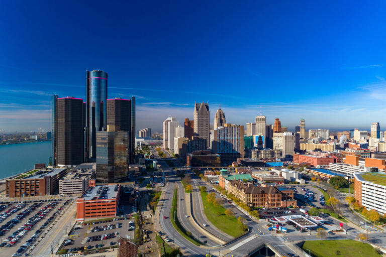 An aerial view of Downtown Detroit skyline with a mostly clear blue sky, and a highway in the foreground and the Detroit river to the left.
