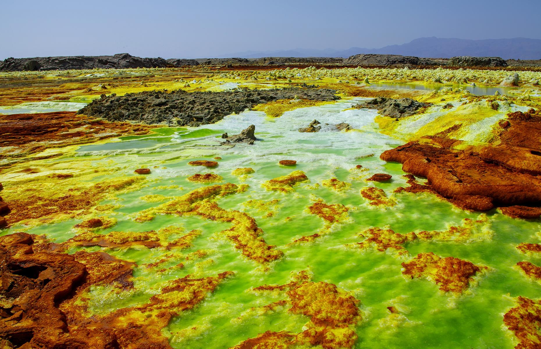Slide 26 of 43: A result of the divergence of three tectonic plates, Ethiopia's Danakil Depression is a sight like no other. Here, steam spits out from openings in the Earth’s crust and chemicals released by the hot springs color the rocky mineral deposits yellow, orange and green. This eerie lunar-like landscape of sulfur springs is also one of the hottest places on Earth.