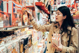 a person standing in front of a shop: A woman browsing the shelves of a store and picking out boxes of holiday sweets.