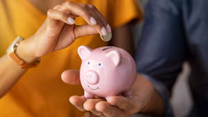 a hand holding a remote control: Close up of man holding pink piggybank while woman putting coin in it.