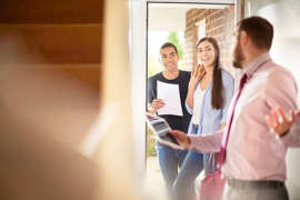 a person standing next to a window: A man and woman looking through the door of a house held open by a realtor.