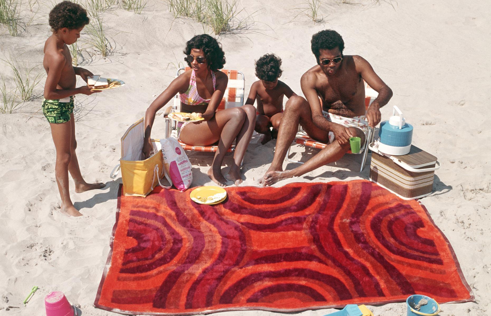 What better place for a picnic than the shortbread-colored shores of America's East Coast? Here a young family snapped in the 1970s begin to lay out a spread on a bright beach towel. Their print swimwear is unmistakably of the decade too.