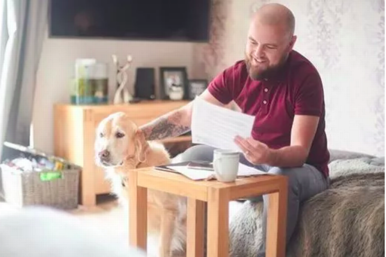 A man is sitting on a sofa smiling as he looks at a letter while a dog is sitting beside him