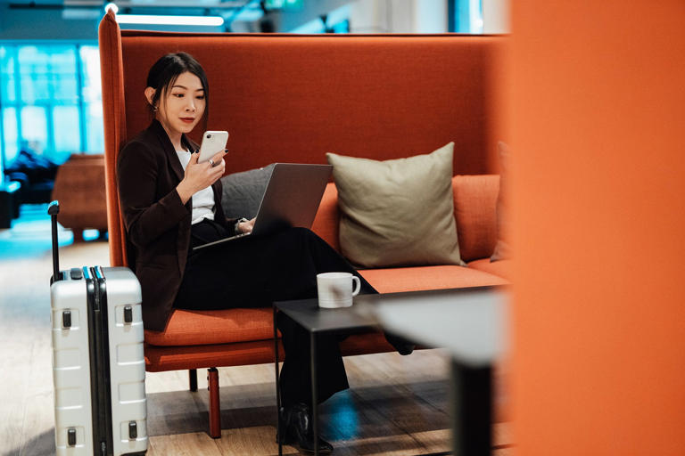 Young Asian business woman working on smartphone and laptop while sitting at airport VIP lounge with suitcase