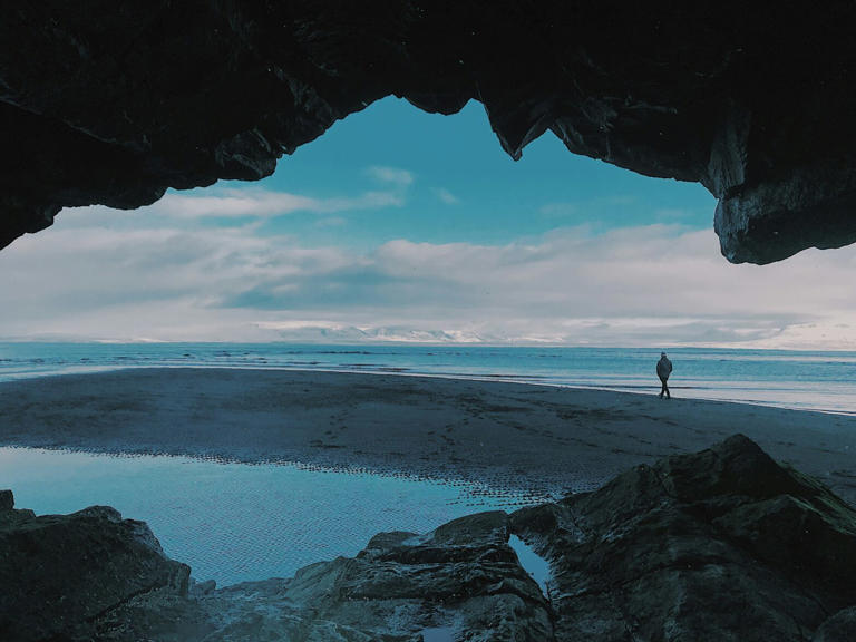 Solo travel is a popular way to explore the world. Check out these top rated places for unaccompanied trips. pictured: a cave opening with a solo traveler standing on the coast of the beach on a cloudy and shady day