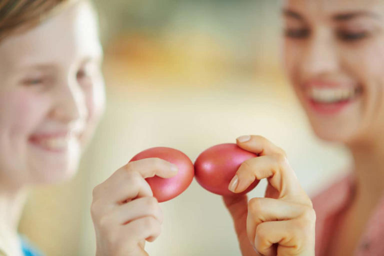 Closeup on happy mother and daughter having egg tapping with red easter eggs in the house in sunny spring day.