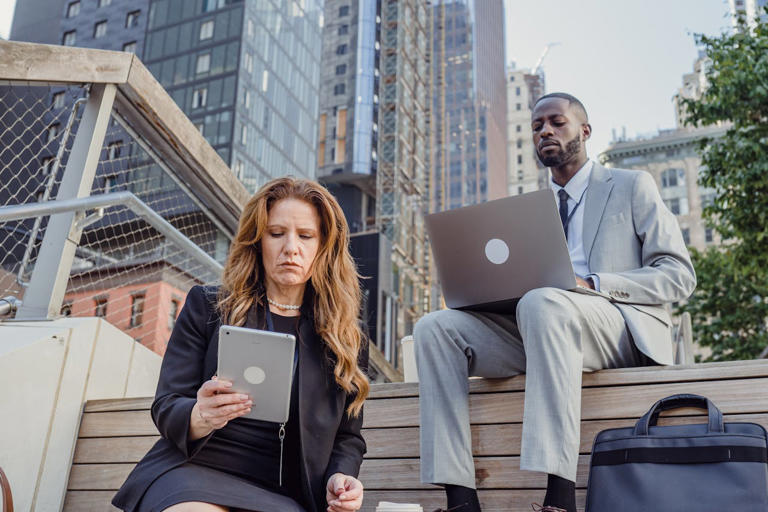 Man and woman sitting outside on steps, outside of a tall building. They are both dressed in business clothes and are looking down seriously at their laptops.
