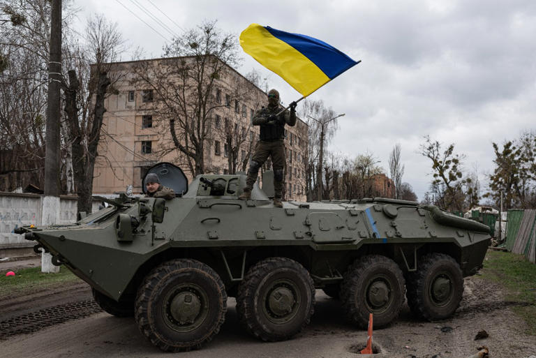 A Ukrainian soldier waves Ukrainian national flag while standing on top of an armoured personnel carrier (APC) on April 8, 2022 in Hostomel, Ukraine. Western countries backing Ukraine need to urgently increase their defense production and dedicate more of their military equipment to Kyiv to prevent the Kremlin triumphing in Ukraine, according to a new analysis.