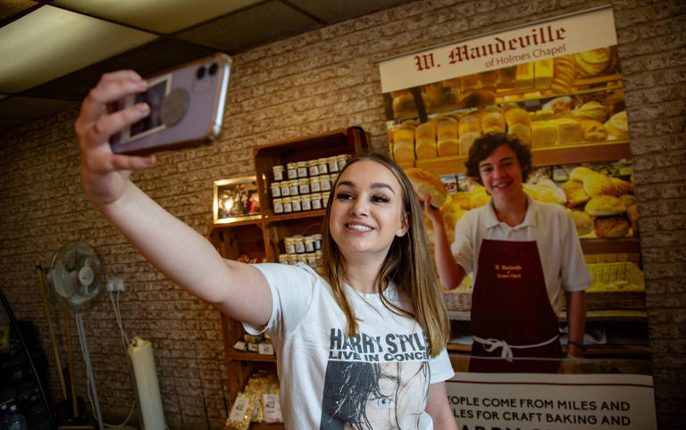 Fan Izzy Hawksworth poses for a selfie in front of a picture of a young Harry Styles proudly displayed at the bakery where he worked - WILLIAM LAILEY/SWNS