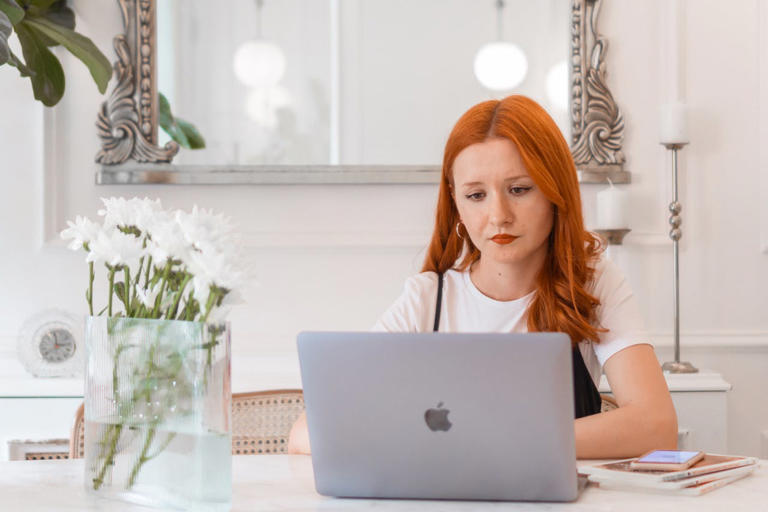Red haired woman sitting at a desk with flowers looking at a laptop.