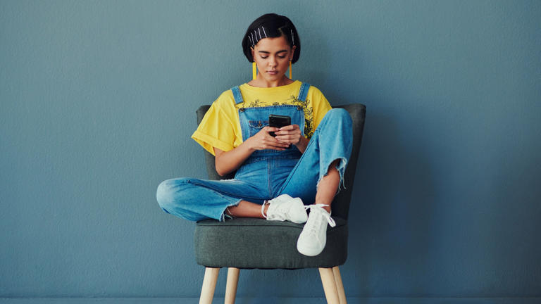 younger woman millennial sits in chair cell mobile smartphone_iStock-1311401106