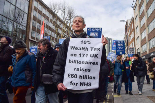 O ativista Peter Tatchell segura um cartaz pedindo imposto sobre a riqueza dos bilionários para financiar o sistema de saúde britânico em 2023 em Londres