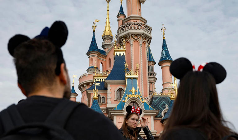 Visitors wearing emblematic Mickey and Minnie Mouse ears look on, in front of the Sleeping Beauty-inspired castle at Disneyland Paris, in Marne-la-Vallee, east of Paris, on October 16, 2023. characters.